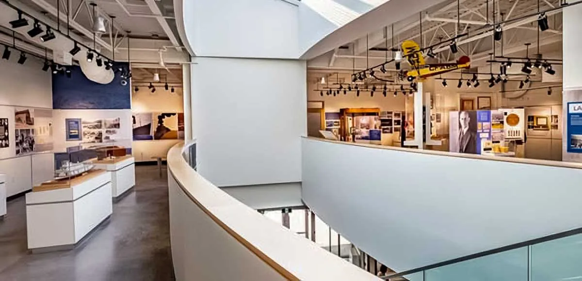Interior view of a modern museum with curved white walls, display cases, photos, and a yellow model airplane hanging from