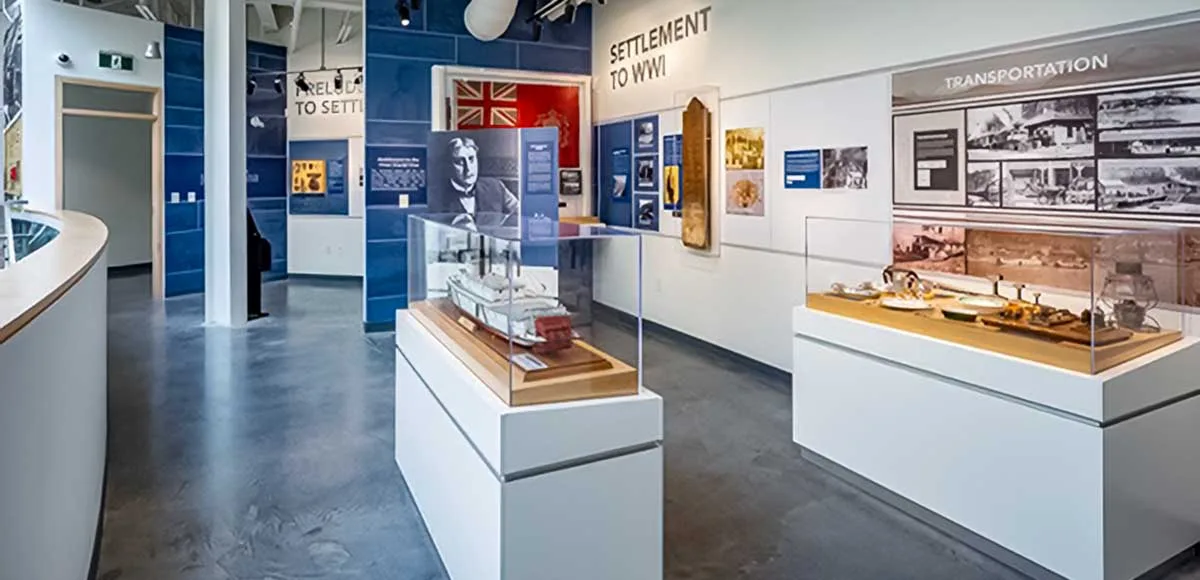 Museum exhibit room with display cases, historical photos, a model ship, and text panels about settlement and transportation.