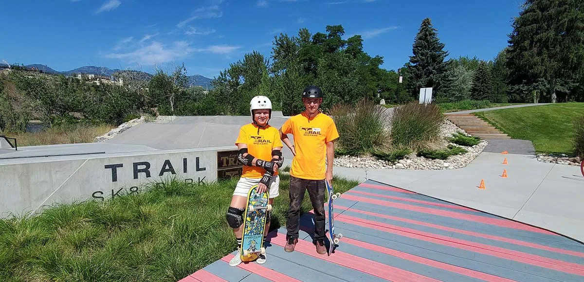 Two people in yellow Trail t-shirts holding skateboards standing on a striped ramp at Trail Skate Park on a sunny day.