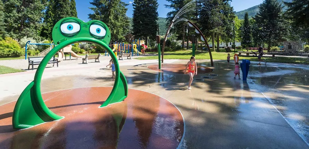 Children playing in a splash pad with frog-shaped water feature and other water sprays in a park with trees and playground.