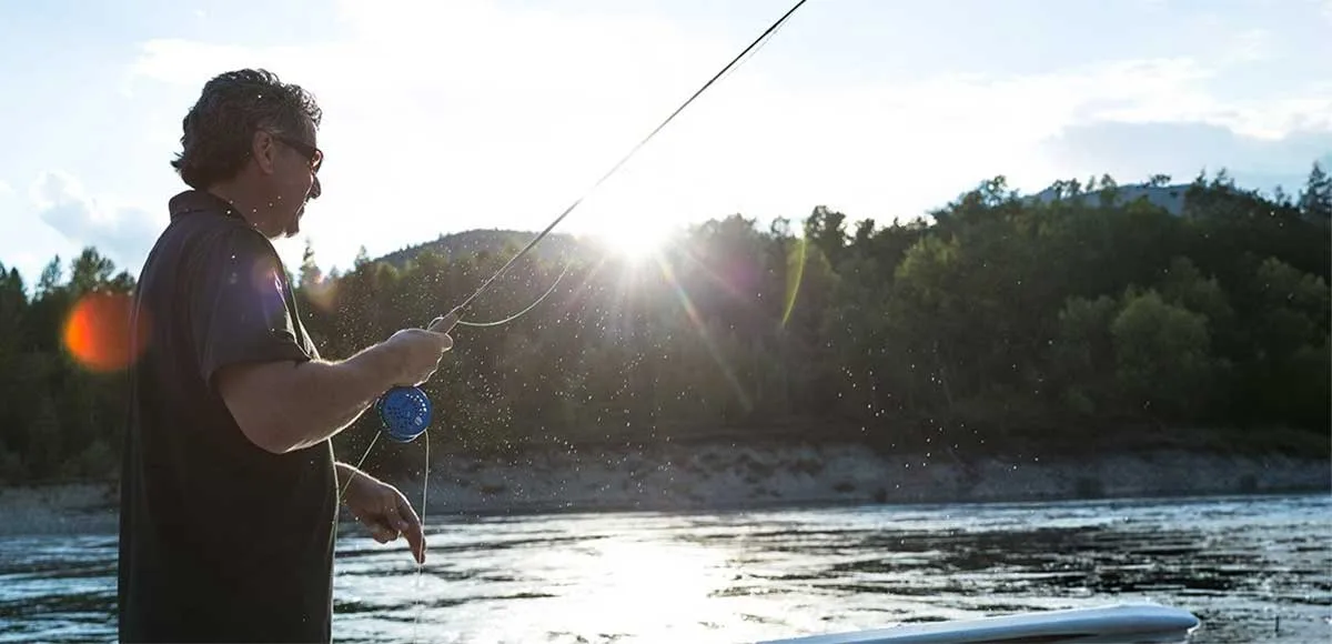 Man fishing with a rod near a river at sunset with trees and hills in the background