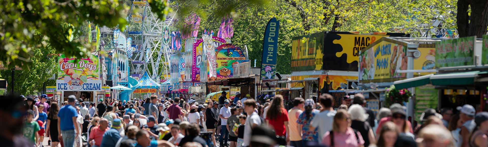Trail BC Crowded outdoor fair with food stalls, colorful signs, and green trees lining the walkway on a sunny day.
