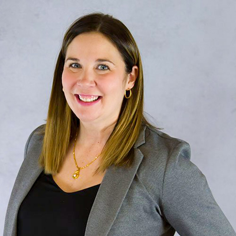 BevBenson2022 Woman with shoulder-length brown hair wearing a gray blazer, black top, gold necklace, and hoop earrings smiling against a