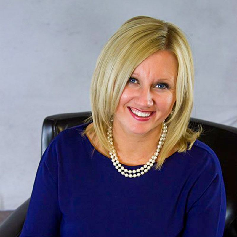 TheaHanson2022 Blonde woman wearing a blue top and pearl necklace, smiling while seated on a black chair against a gray background.