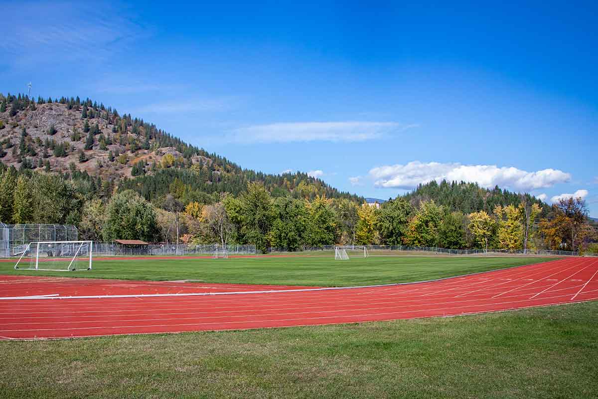 Outdoor sports field with soccer goals surrounded by a red running track, trees, and hills under a blue sky.