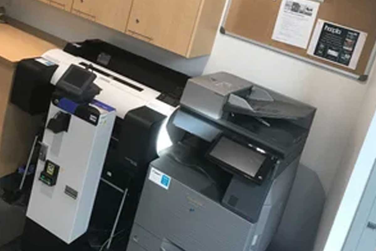 Printing Services Two large multifunction printers/copiers in a corner under wooden cabinets and a bulletin board on the wall above.
