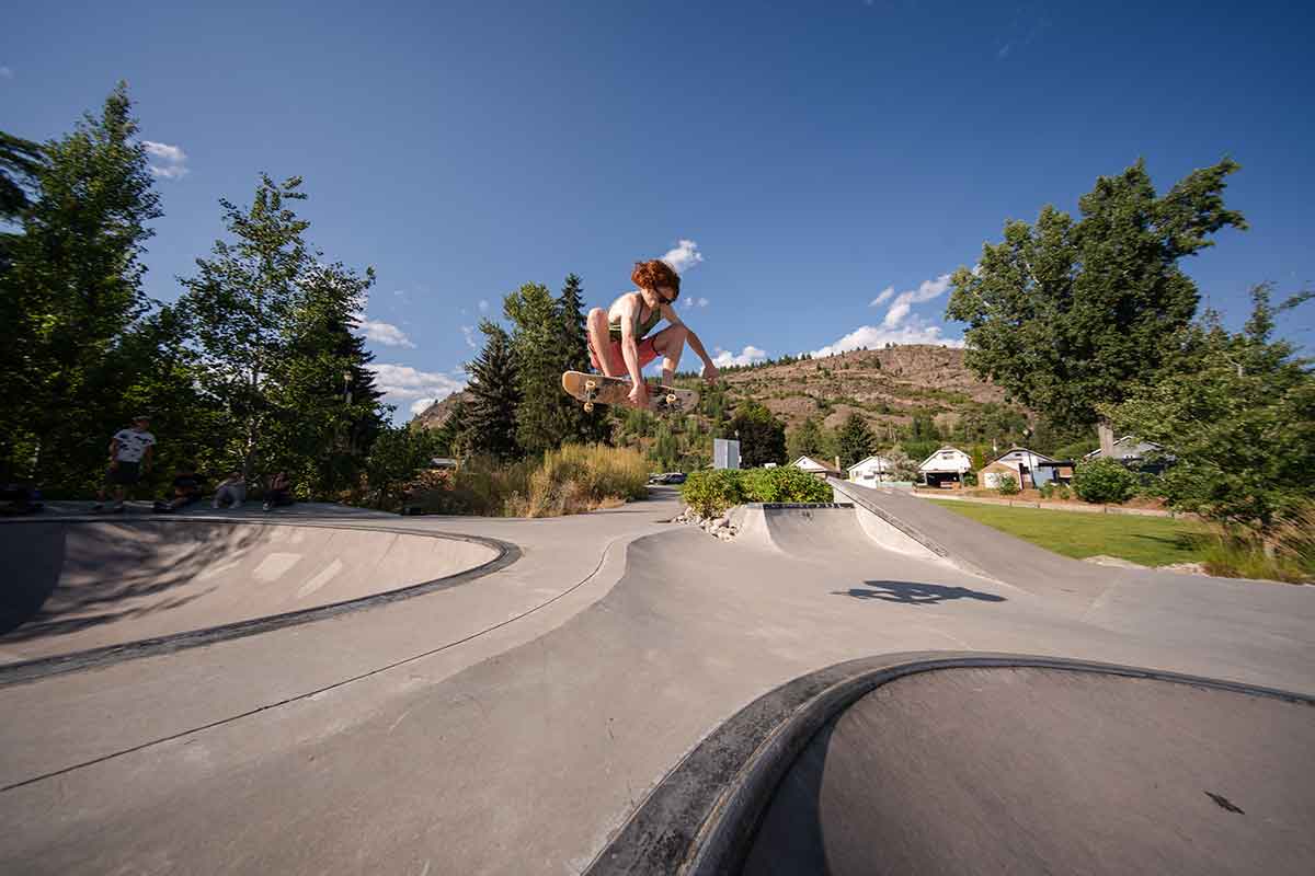 Skateboarder mid-air performing a trick at a concrete skate park with trees and houses in the background.