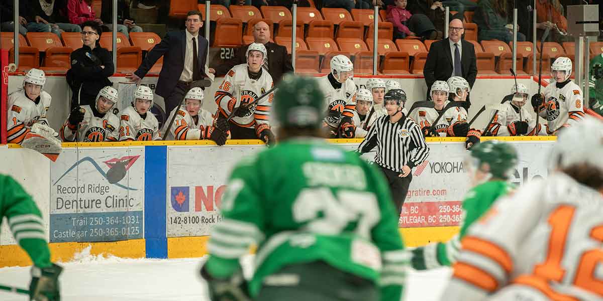 Hockey players in white and orange jerseys sit on the bench with coaches standing behind during a game.