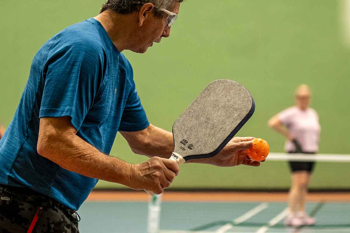 Man in blue shirt holding a pickleball paddle and orange ball on an indoor court with a woman in the background