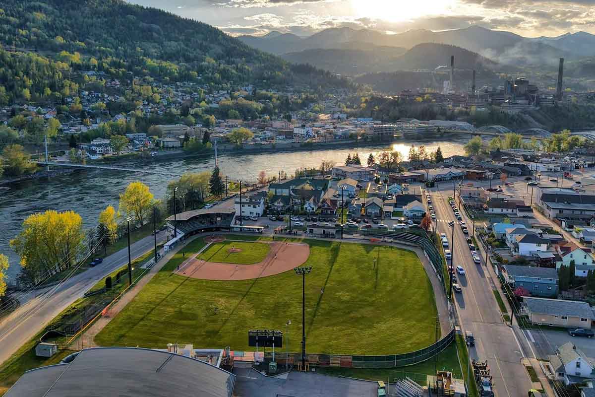 Aerial view of a baseball field surrounded by houses, a river, hills, and a setting sun in the background.
