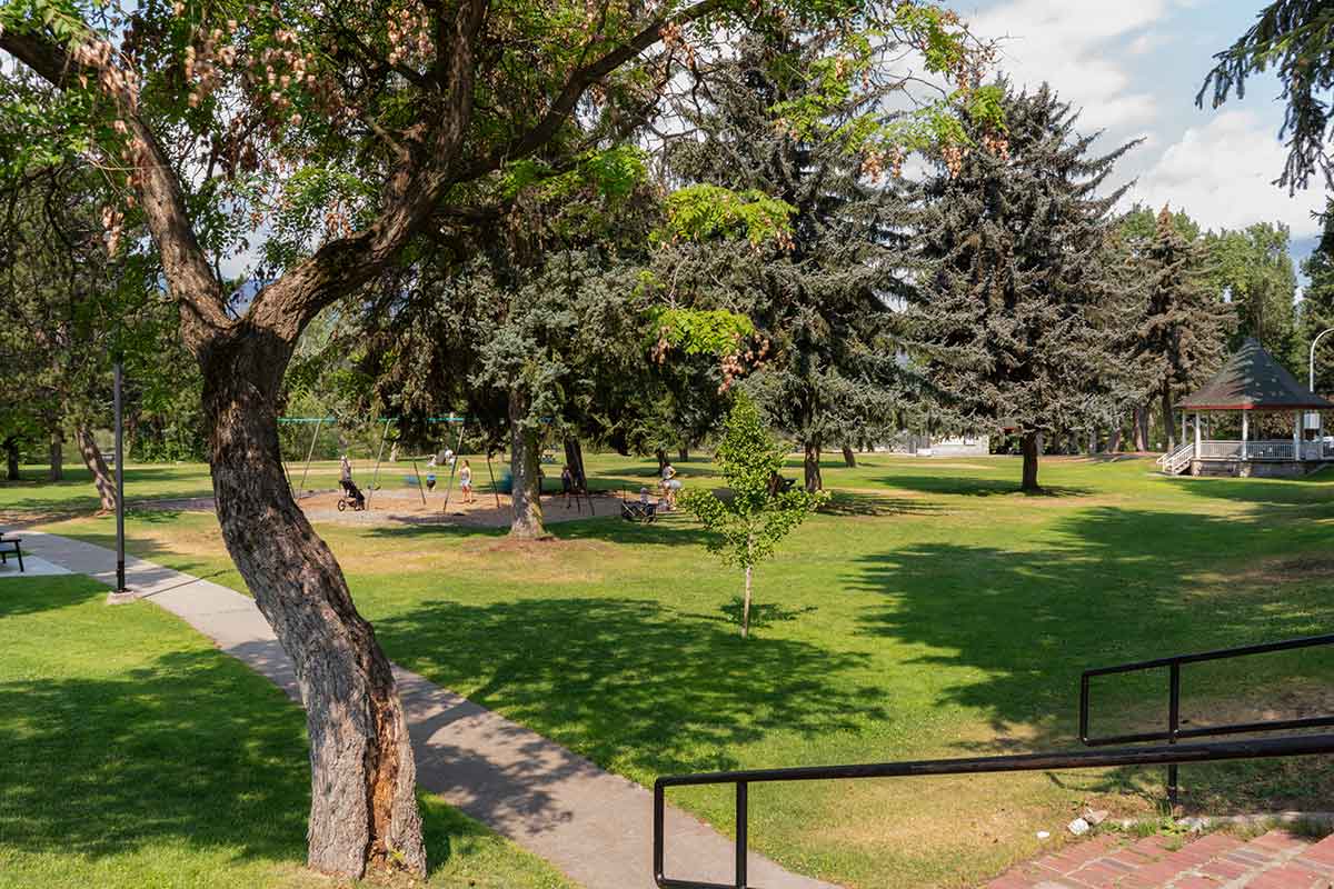 Park scene with large trees, a playground with swings, a paved path, and a gazebo in the background on a sunny day.