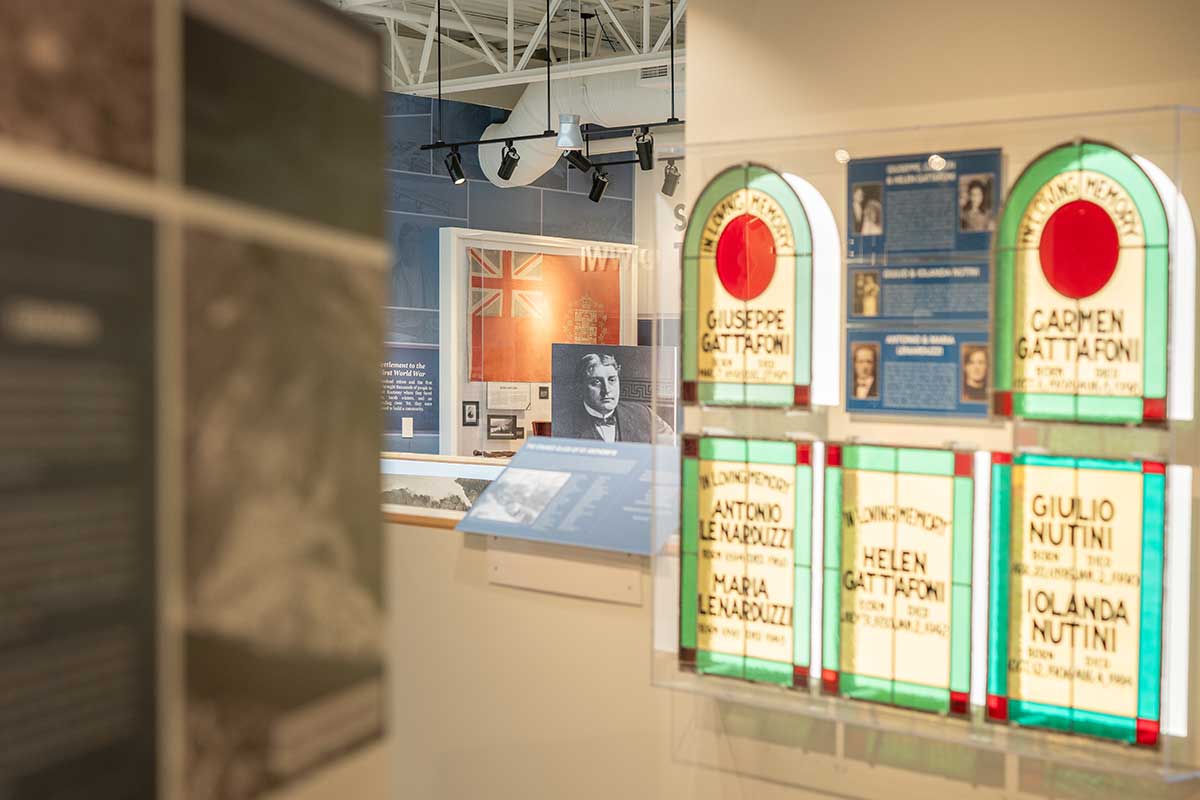 history2 Stained glass panels with names and dates displayed in a museum exhibit with historical photos and flags in the background.