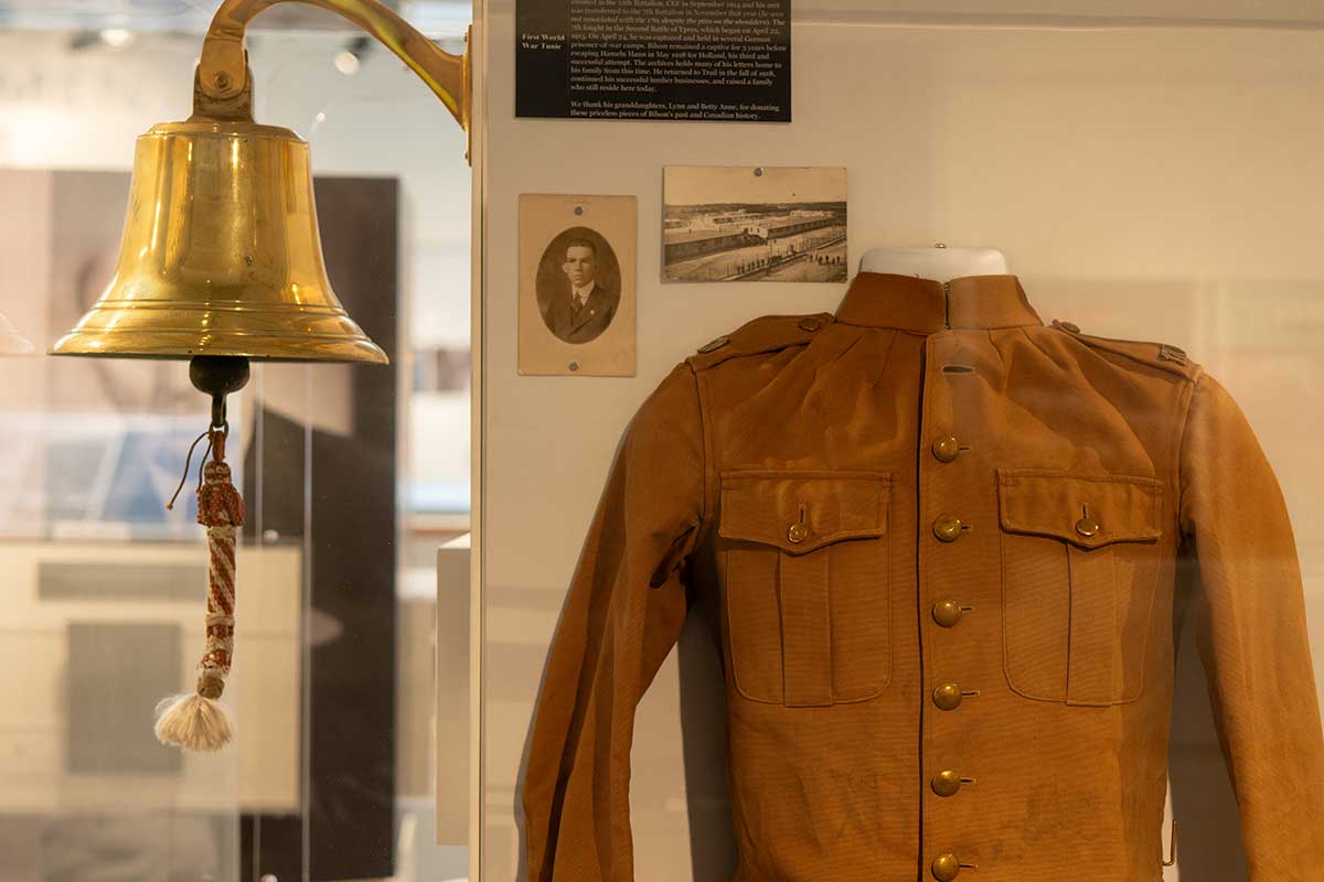 history3 Brass bell with rope pull next to a brown military jacket displayed in a museum exhibit case with photos and text.