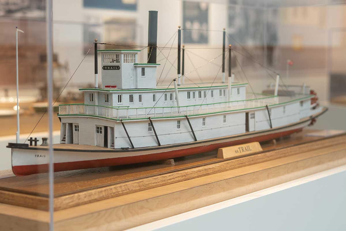 history4 Model of a white and red paddle steamer boat named Trail displayed in a glass case on a wooden base.