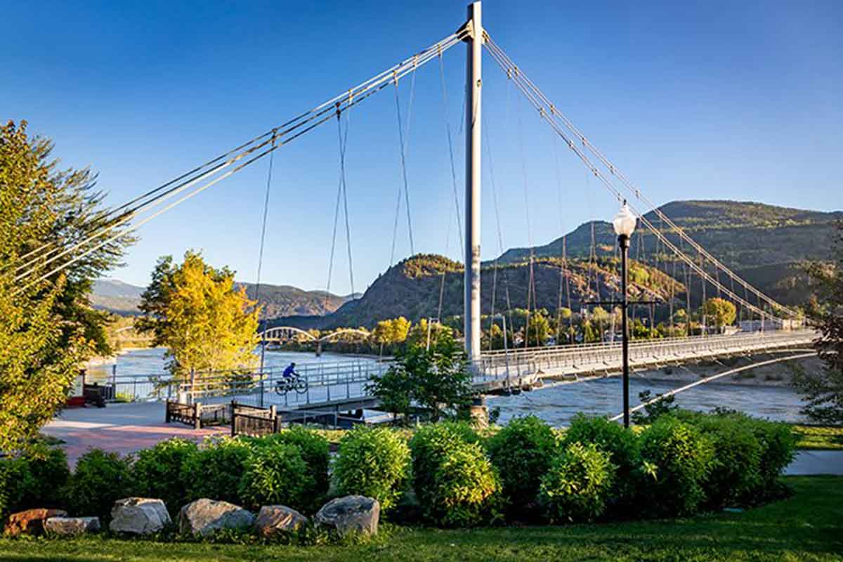 Suspension bridge over river with cyclist, green bushes, trees, mountains, and clear blue sky in background