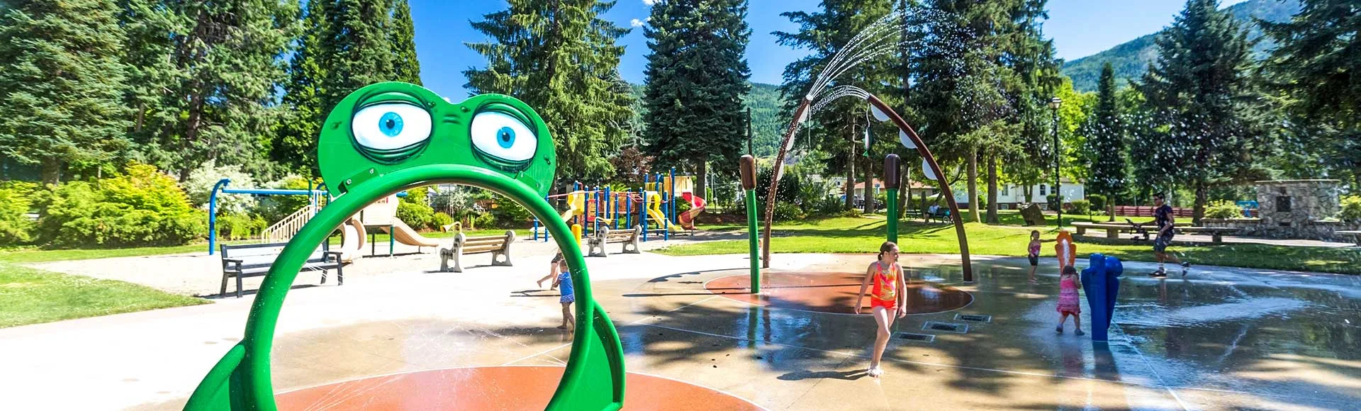tbar_parks2 Children playing in a splash pad area with a green frog water feature and playground equipment surrounded by trees and