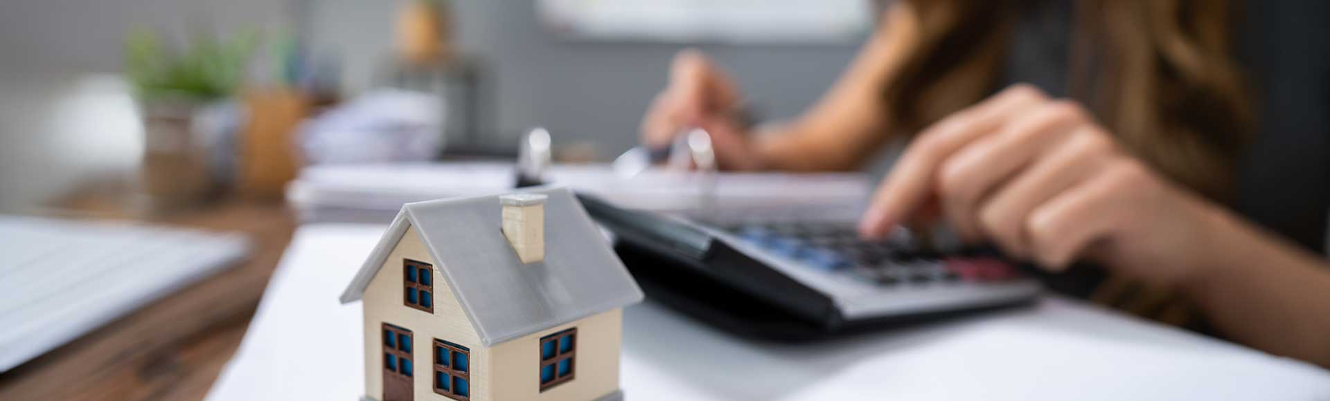 Small model house on desk with person using calculator and writing on paper in background