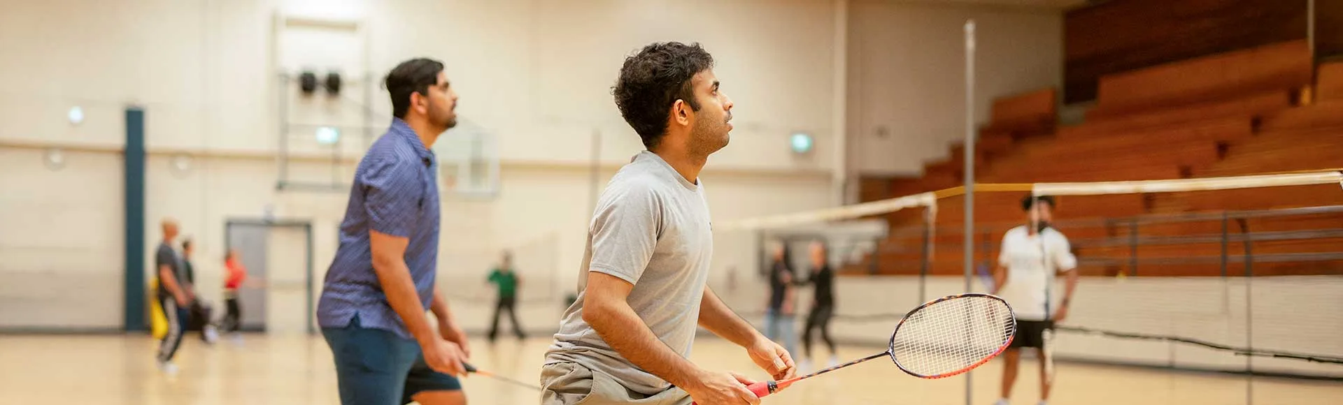 tbar_recreation3 Two men playing badminton in an indoor gymnasium with other players and wooden bleachers in the background
