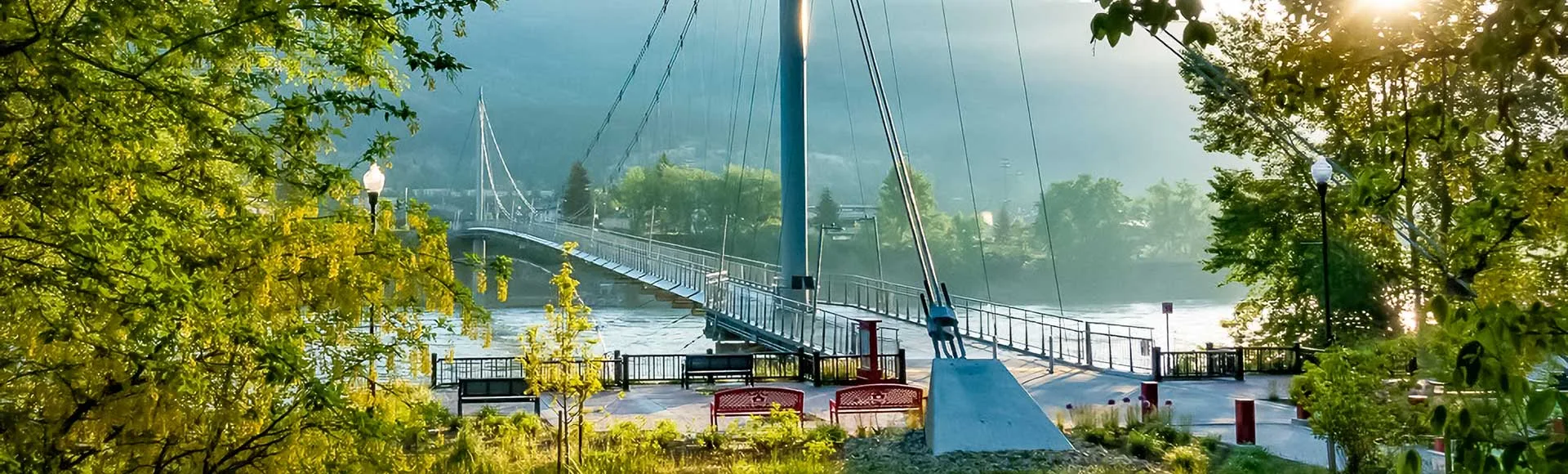 tbar_skywalk Suspension bridge over river surrounded by green trees and park benches in soft sunlight.