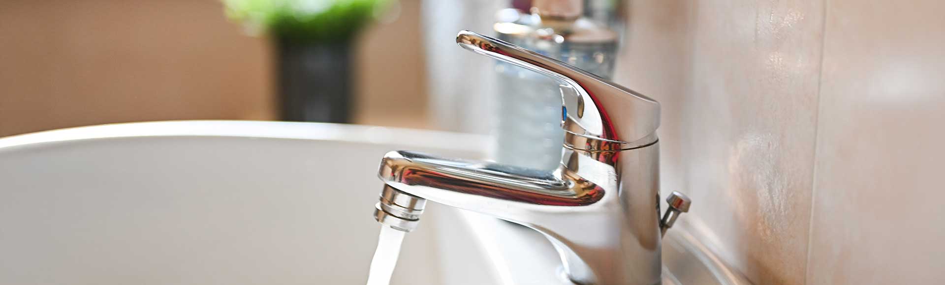 Chrome faucet running water into white bathtub with beige tiled wall and blurred background objects.