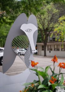 PIcasso resized Abstract metal sculpture resembling a stylized human face with orange flowers in the foreground and trees in the background