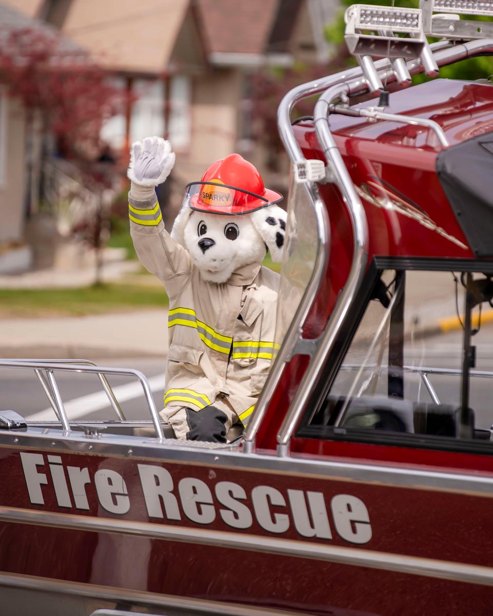 SCD Gallery_04 Person in Sparky the Fire Dog costume waving from a red Fire Rescue vehicle during a parade or event.