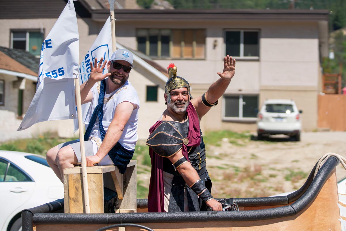 SCD Gallery_05 Two men waving from a parade float, one in Roman soldier costume and the other in casual clothes with sunglasses and a cap.