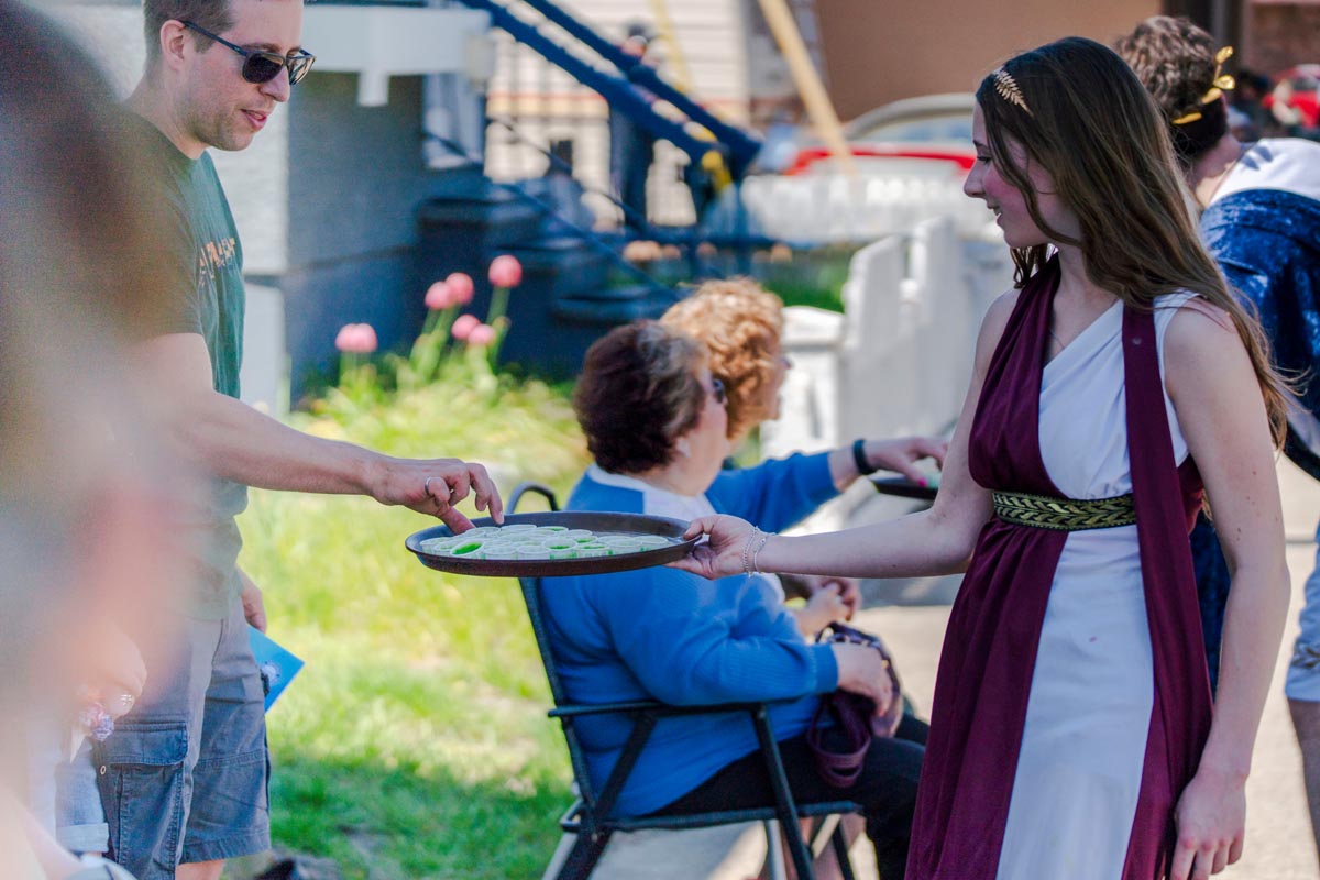 SCD Gallery_08 Young woman in toga-style dress offers tray of cookies to man outdoors, with seated people and house in background
