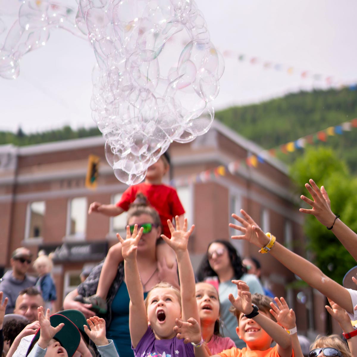 SCD Gallery_09 Children and adults reaching up excitedly toward large floating soap bubbles at an outdoor event in a town square.
