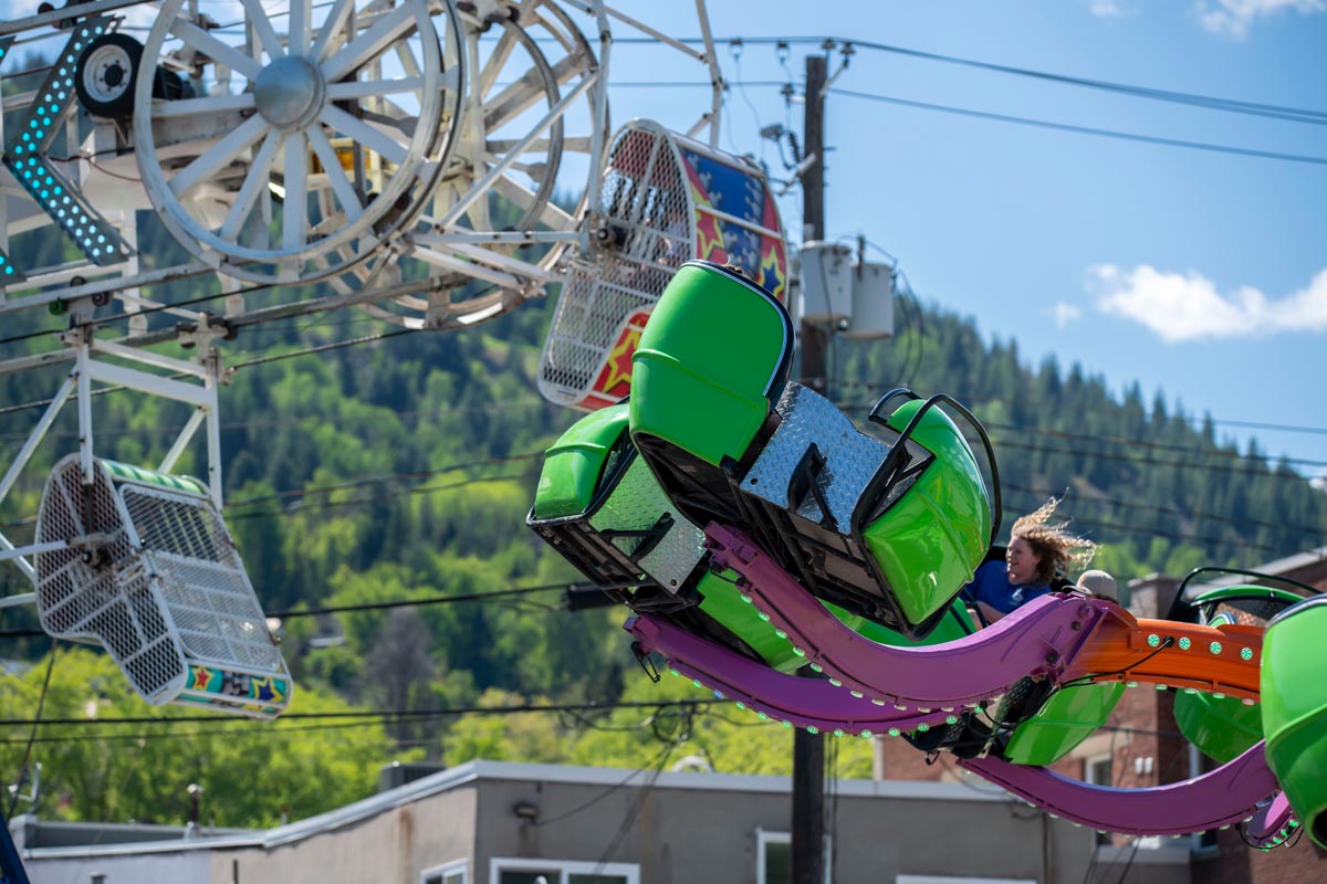SCD Gallery_12 Green and purple amusement ride with a person riding, Ferris wheel and forested hills in the background under blue sky