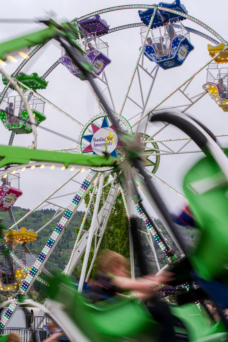 SCD Gallery_14 Colorful Ferris wheel with people in seats and blurred green swing ride in foreground at outdoor amusement park.
