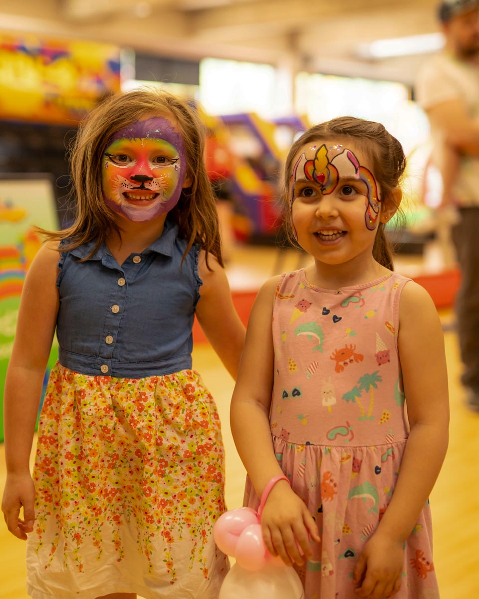 SCD Gallery_18 Two young girls with colorful face paint, one holding a pink balloon animal, smiling indoors at an event.