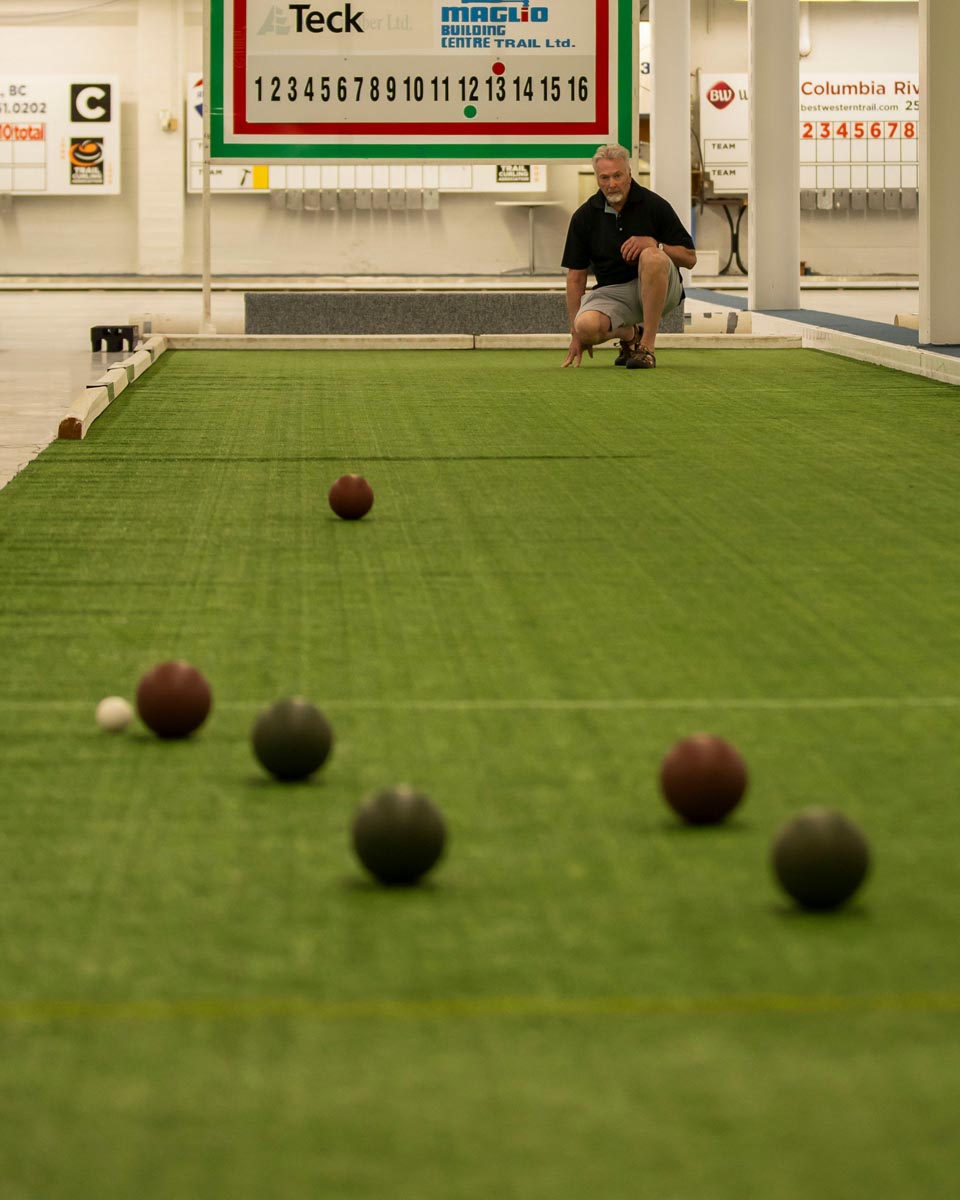 SCD Gallery_19 Man crouching on indoor bocce court aiming to roll a ball toward several balls near the foreground on green turf.
