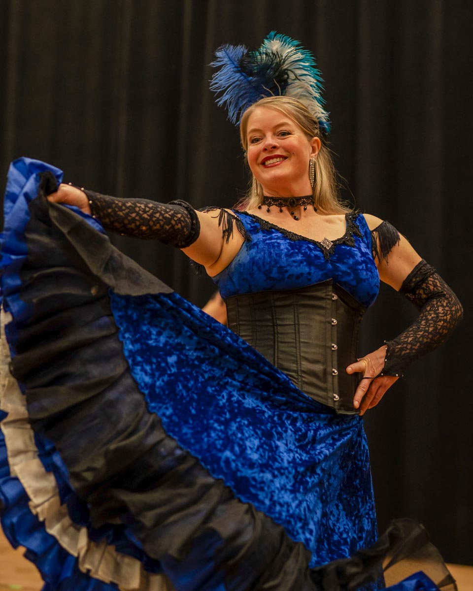 SCD Gallery_21 Woman in blue velvet dress with black corset and feathered headpiece dancing with skirt lifted against black background