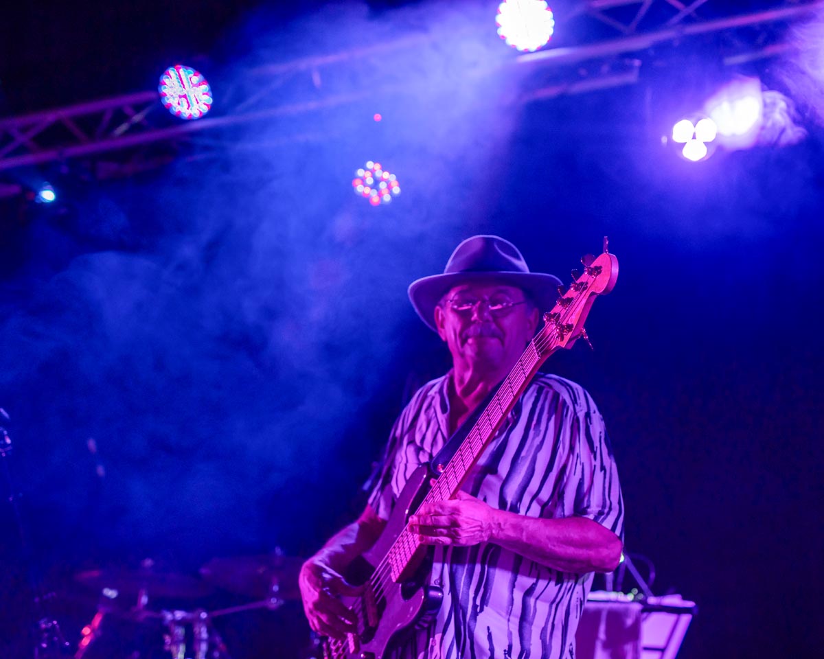 SCD Gallery_24 Man wearing a hat playing electric bass guitar on stage with purple and blue lighting and smoke effects