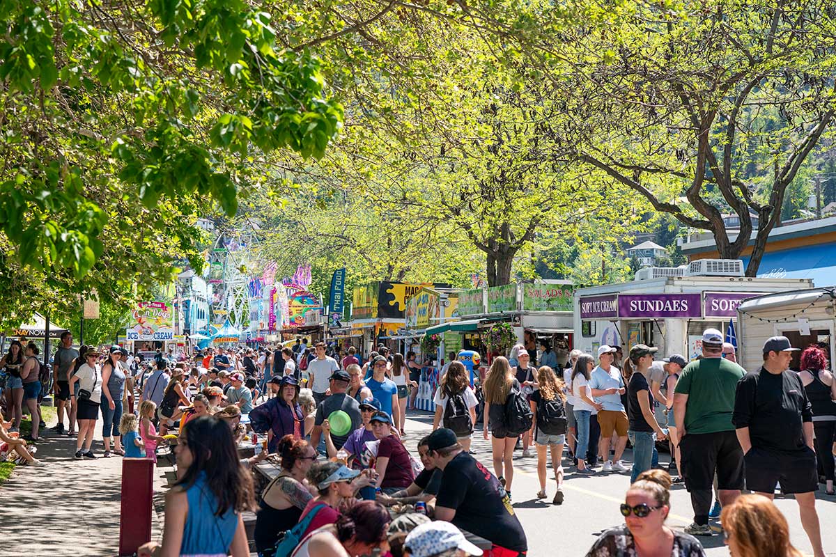 Crowd of people walking and sitting along a tree-lined street with food stalls and carnival rides in the background.