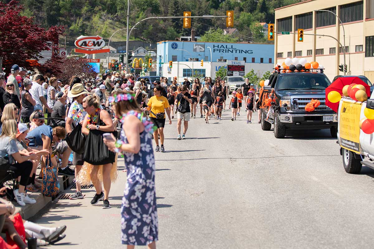 Crowd watching a parade on a sunny street with decorated trucks and people walking in costumes and casual clothes.