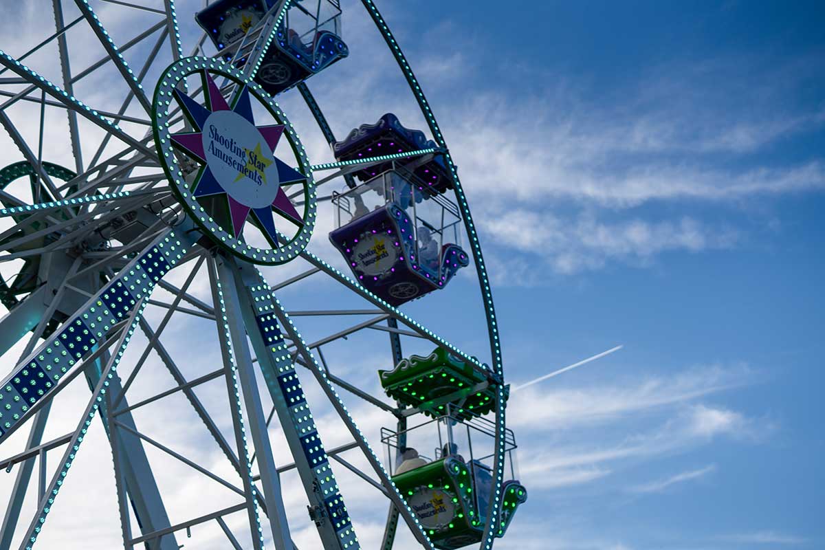 Ferris wheel with colorful lit gondolas against a blue sky with wispy clouds and a contrail.