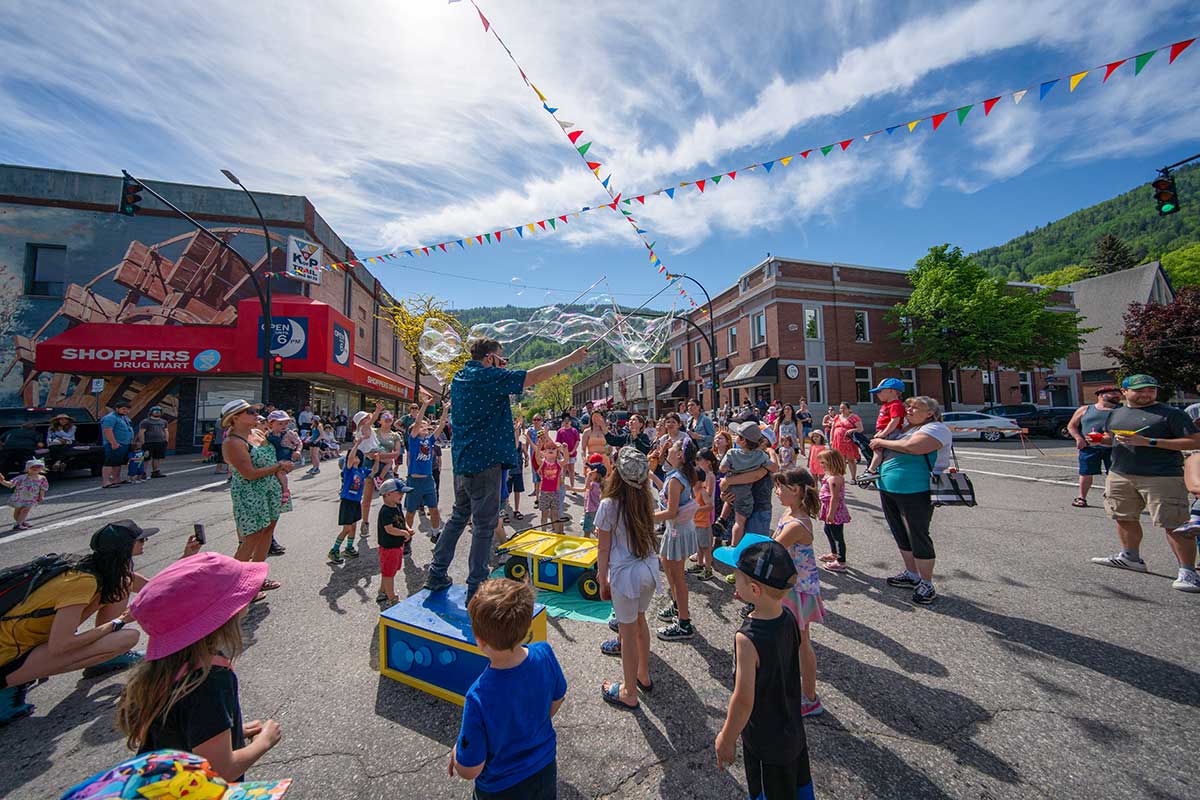 Crowd of children and adults watching a man making large soap bubbles on a sunny street decorated with colorful flags.