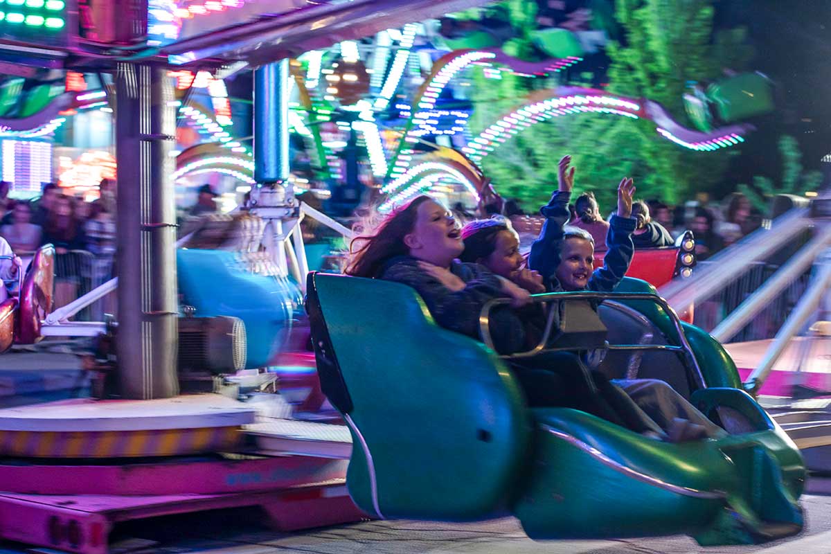 Three children smiling and raising hands while riding a spinning amusement park ride at night with colorful lights.