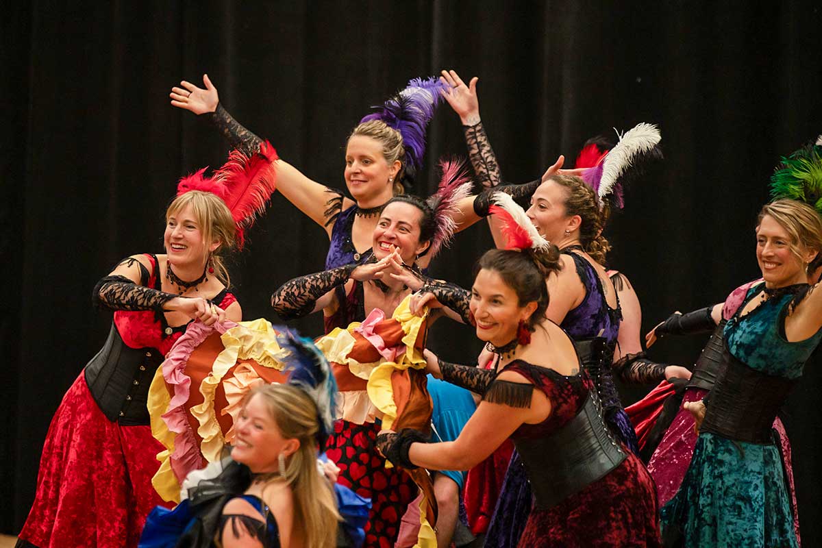 Seven women in colorful can-can style costumes with feathered headpieces dancing against a black background